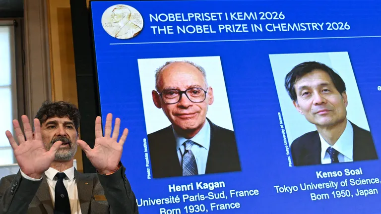 A man stands with his hands up in front of a screen displaying two men in suits against a blue background.
